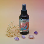 The Coconut Kiss Argan Body Oil surrounded by crystals and a dried flower on a pink background.