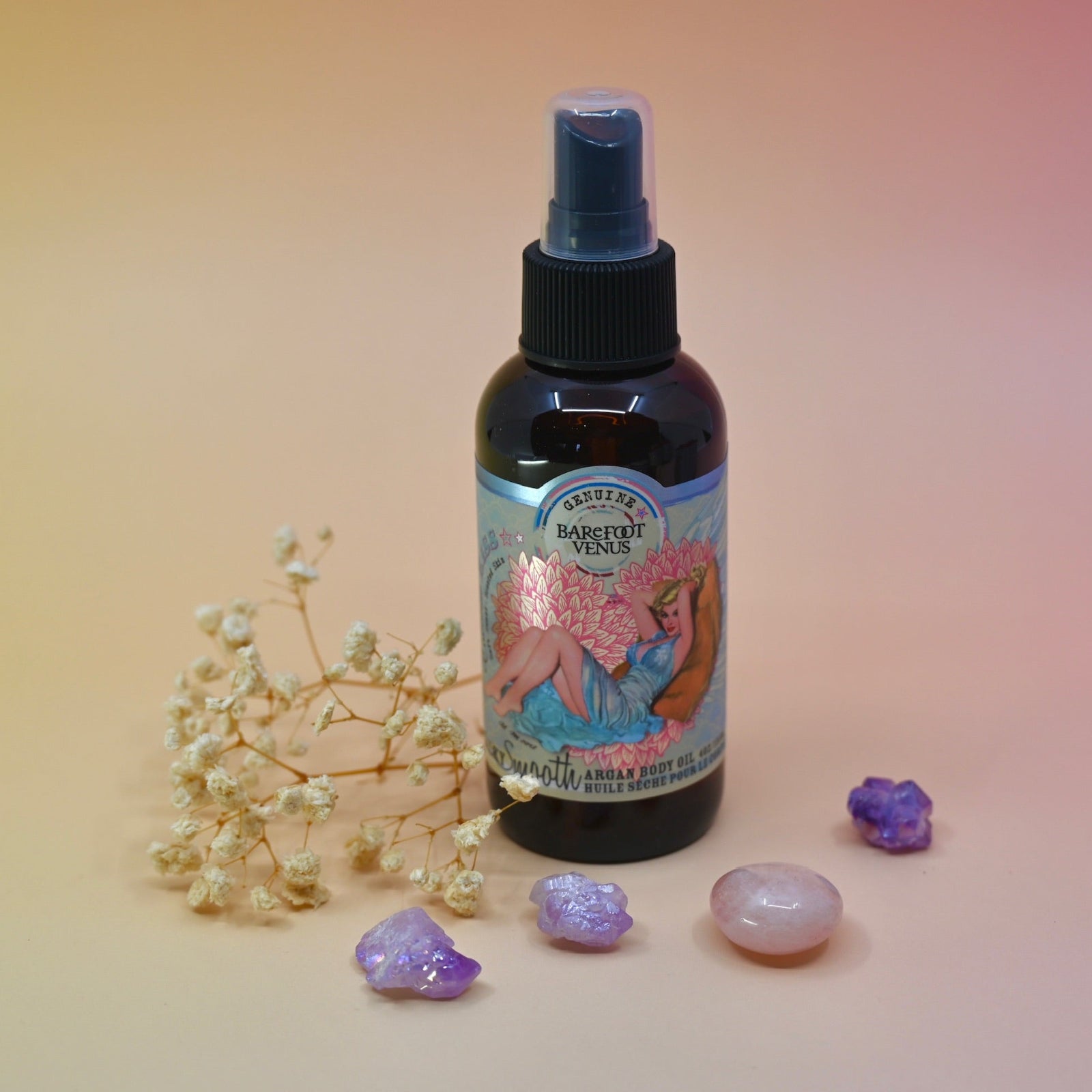 The Coconut Kiss Argan Body Oil surrounded by crystals and a dried flower on a pink background.