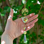 The citrine Tree of Life Crystal Suncatcher has someone's hand behind it with greenery in the background.
