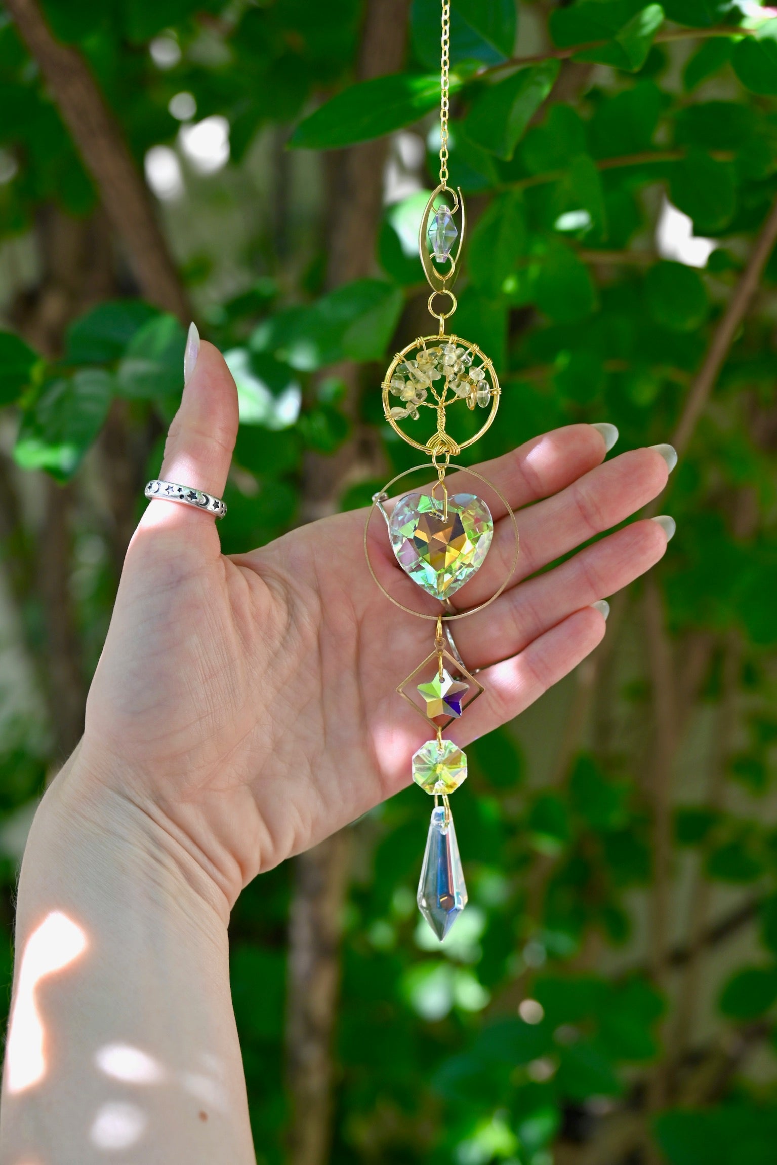 The citrine Tree of Life Crystal Suncatcher has someone's hand behind it with greenery in the background.