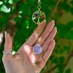 The chakra Tree of Life Crystal Suncatcher has someone's hand behind it with greenery in the background.