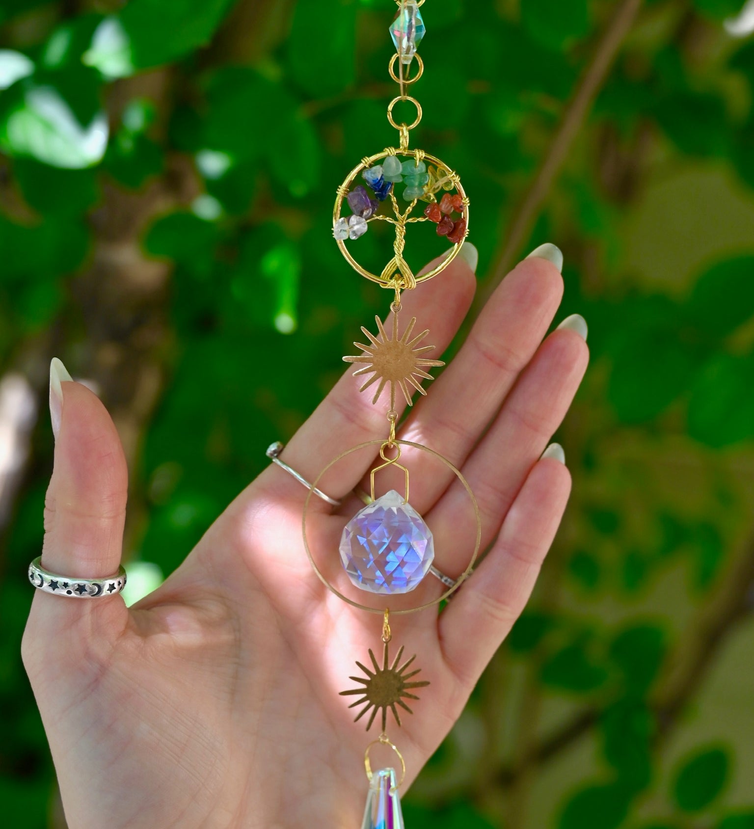 The chakra Tree of Life Crystal Suncatcher has someone's hand behind it with greenery in the background.