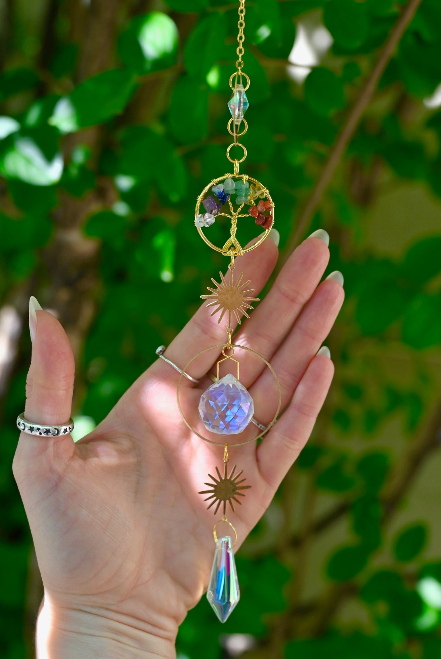 The chakra Tree of Life Crystal Suncatcher has someone's hand behind it with greenery in the background.
