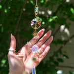 The chakra Tree of Life Crystal Suncatcher has someone's hand behind it with greenery in the background.