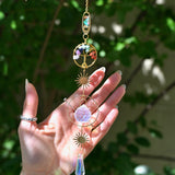 The chakra Tree of Life Crystal Suncatcher has someone's hand behind it with greenery in the background.
