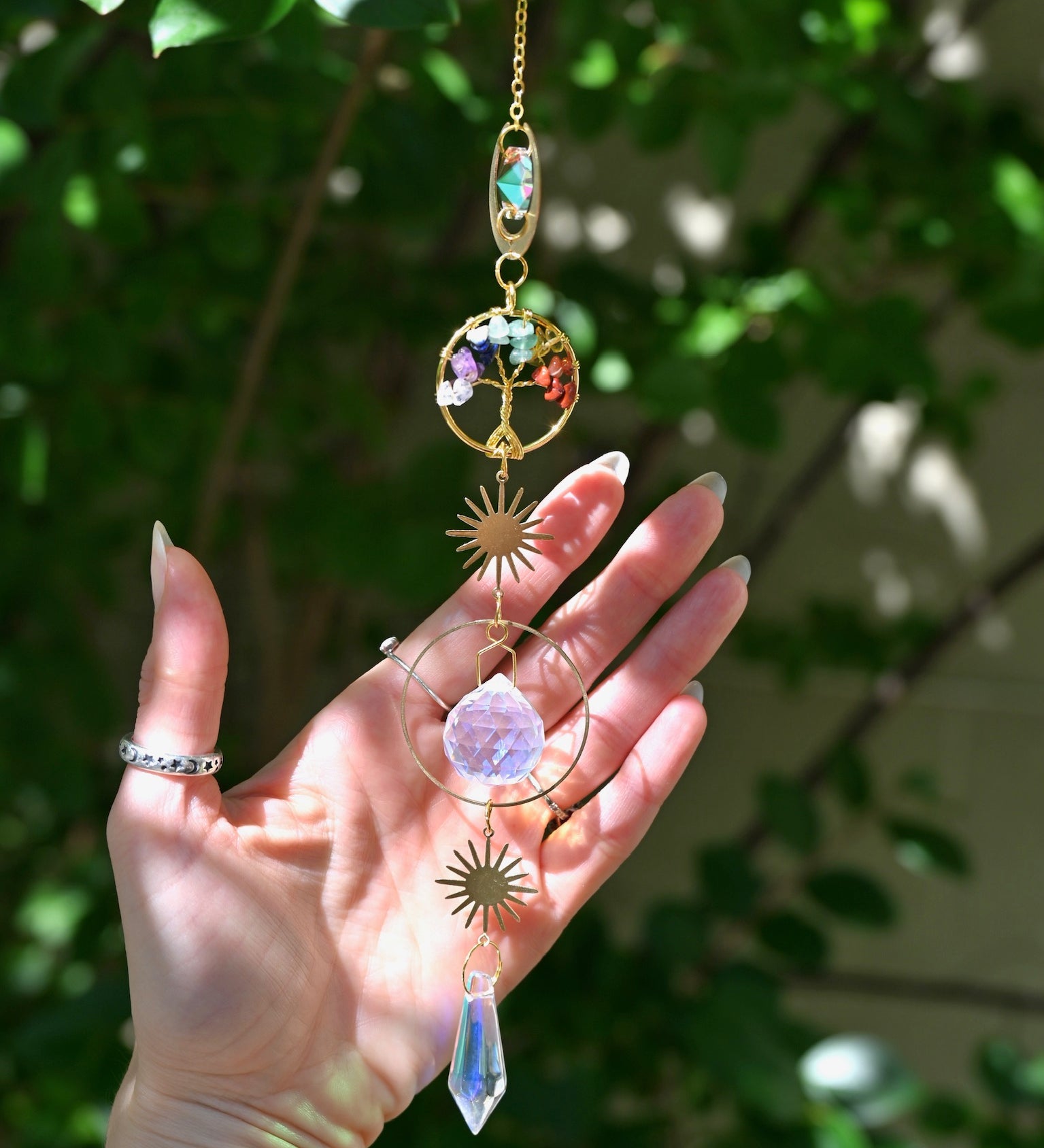 The chakra Tree of Life Crystal Suncatcher has someone's hand behind it with greenery in the background.