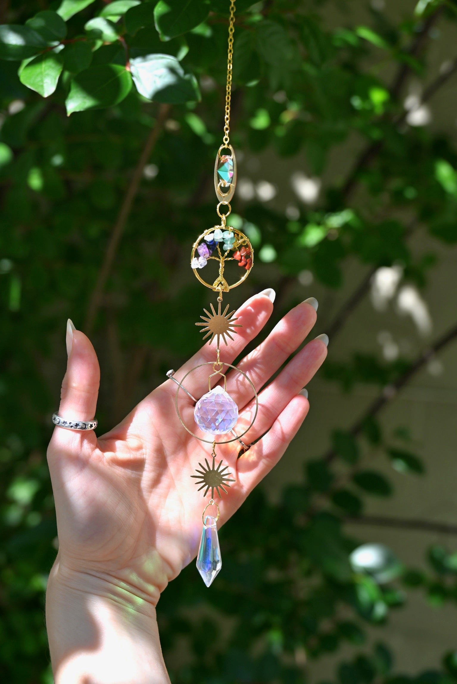 The chakra Tree of Life Crystal Suncatcher has someone's hand behind it with greenery in the background.