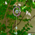 The Amethyst Gemstone Suncatcher in front of a cream-colored brick wall and greenery.