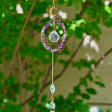 The Amethyst Gemstone Suncatcher in front of a cream-colored brick wall and greenery.