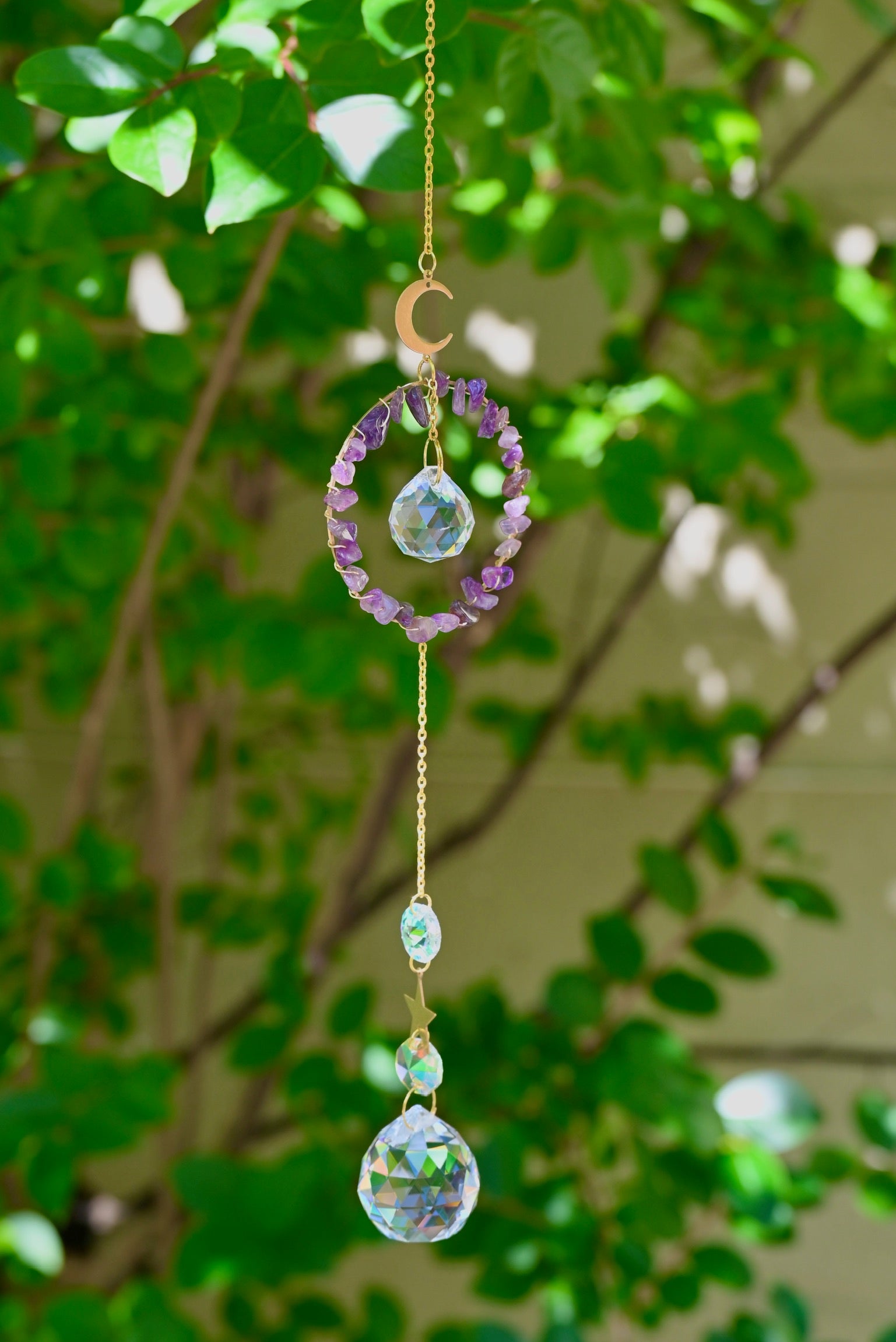 The Amethyst Gemstone Suncatcher in front of a cream-colored brick wall and greenery.