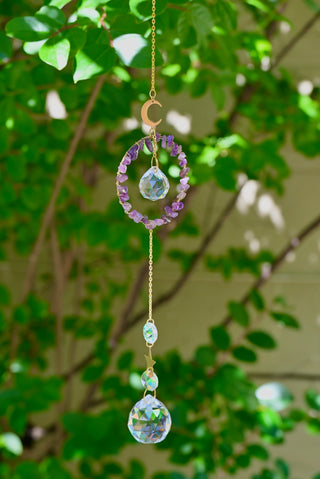 The Amethyst Gemstone Suncatcher in front of a cream-colored brick wall and greenery.