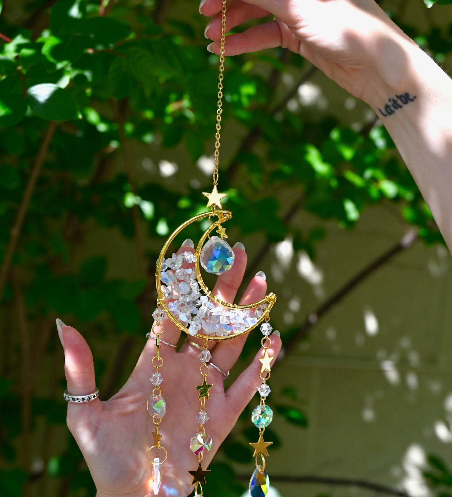 Someone holding the Opalite Crystal Moon Suncatcher with greenery in the background.