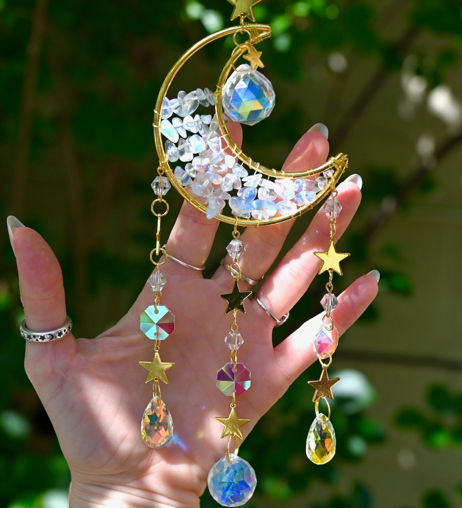 Someone holding the Opalite Crystal Moon Suncatcher with greenery in the background.