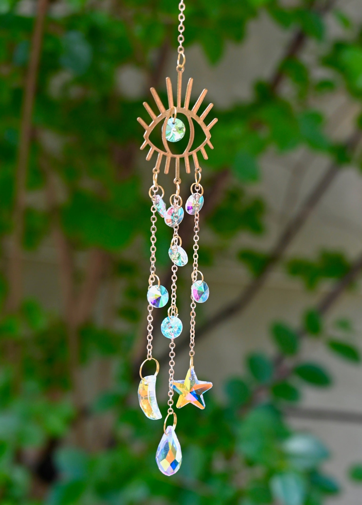 The evil eye  Fairy Lights Suncatcher in front of a cream-colored brick wall with greenery.