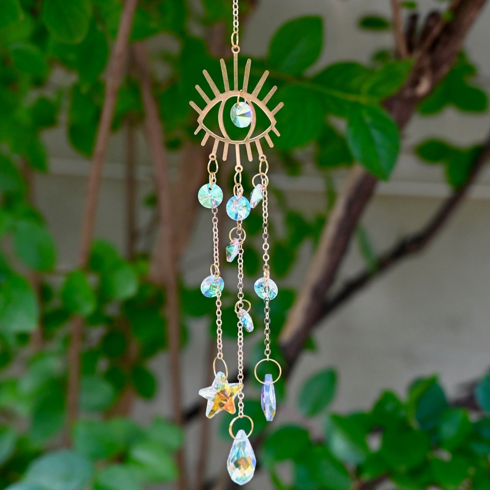 The evil eye  Fairy Lights Suncatcher in front of a cream-colored brick wall with greenery.