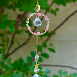 The Watermelon Quartz Gemstone Suncatcher in front of a cream-colored brick wall and greenery.