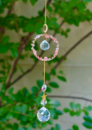The Watermelon Quartz Gemstone Suncatcher in front of a cream-colored brick wall and greenery.