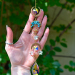 The turquoise Tree of Life Crystal Suncatcher has someone's hand behind it with greenery in the background.