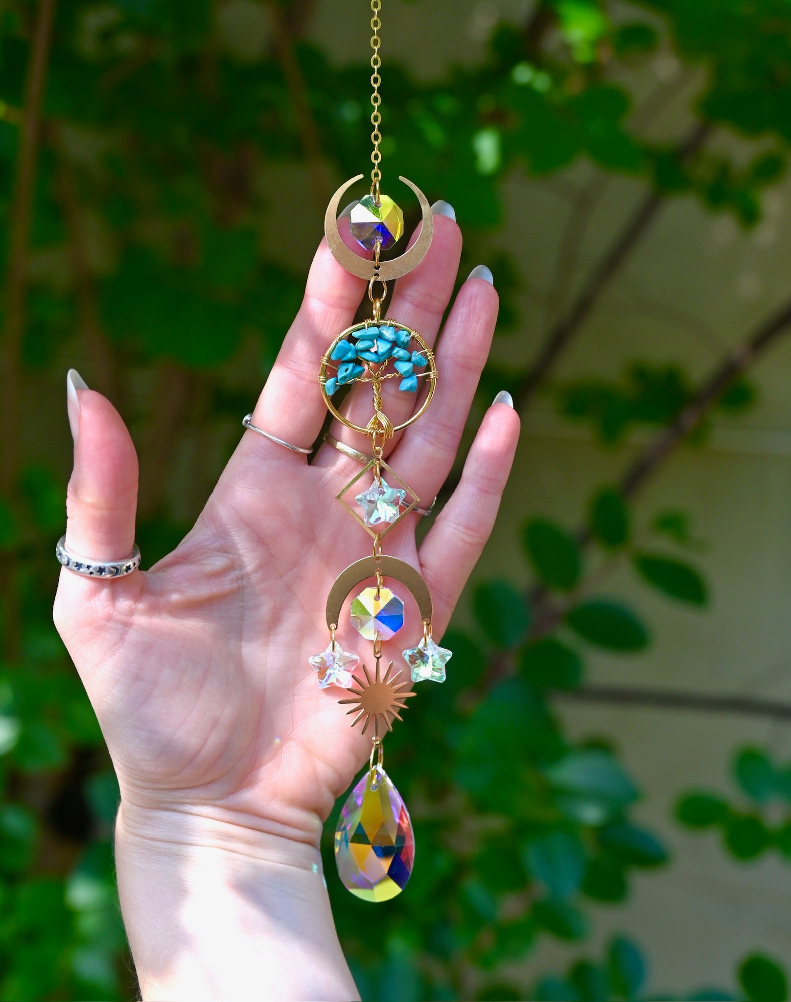 The turquoise Tree of Life Crystal Suncatcher has someone's hand behind it with greenery in the background.