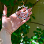 The Aquamarine Gemstone Suncatcher in front of a cream-colored brick wall and greenery.