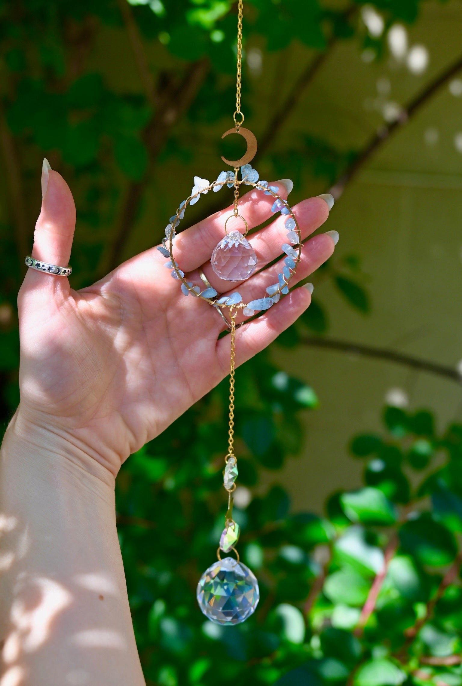 The Aquamarine Gemstone Suncatcher in front of a cream-colored brick wall and greenery.