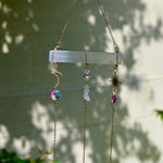 The Selenite Crystal Suncatcher in front of a white brick wall with leaves in the background.