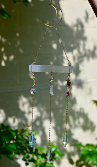 The Selenite Crystal Suncatcher in front of a white brick wall with leaves in the background.