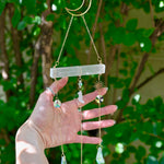 The Selenite Crystal Suncatcher in front of greenery with someone holding the gold chains.