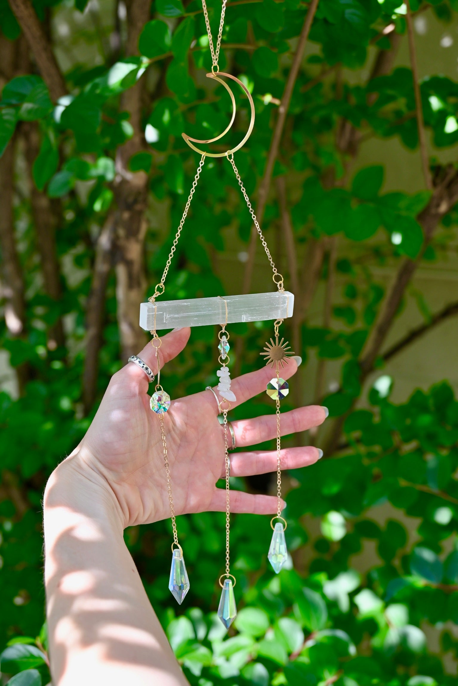 The Selenite Crystal Suncatcher in front of greenery with someone holding the gold chains.