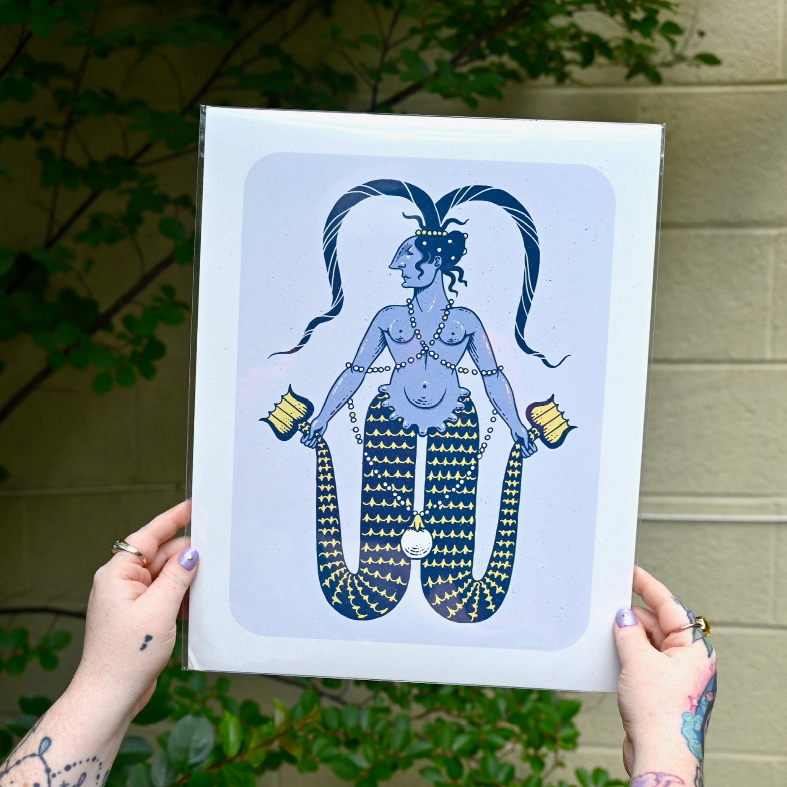 The Twin-Tailed Mermaid Print being held in front of a cream-colored brick wall with greenery.