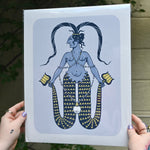 The Twin-Tailed Mermaid Print being held in front of a cream-colored brick wall with greenery.