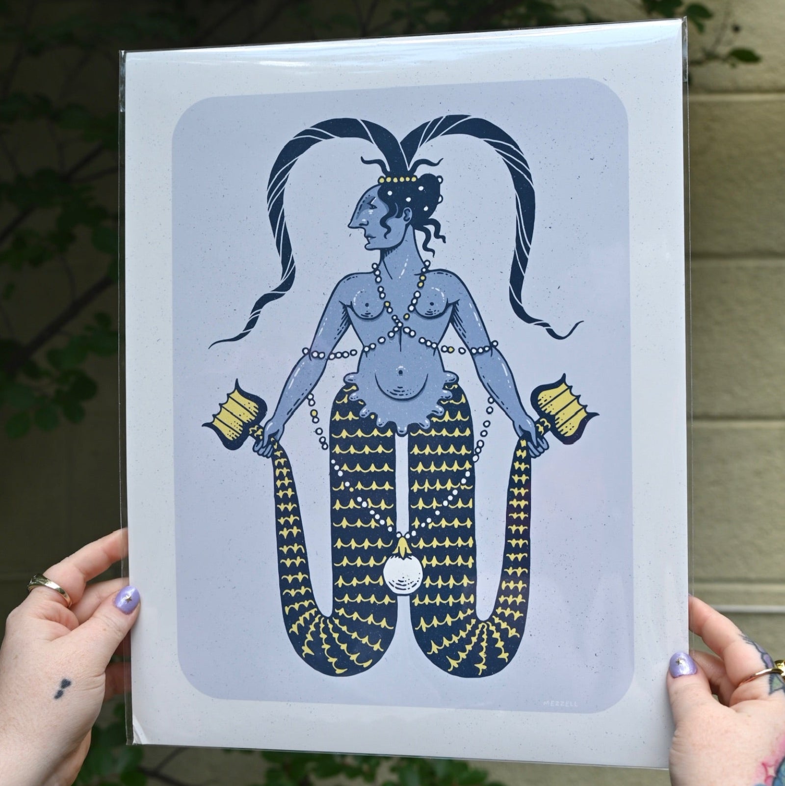 The Twin-Tailed Mermaid Print being held in front of a cream-colored brick wall with greenery.