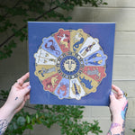 The Mystical Zodiac Print being held in front of a cream-colored wall with greenery.