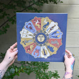 The Mystical Zodiac Print being held in front of a cream-colored wall with greenery.