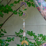 Someone holding the Chakra Gemstone Suncatcher with Amethyst, Citrine, and Quartz in front of a cream-colored brick wall with greenery.