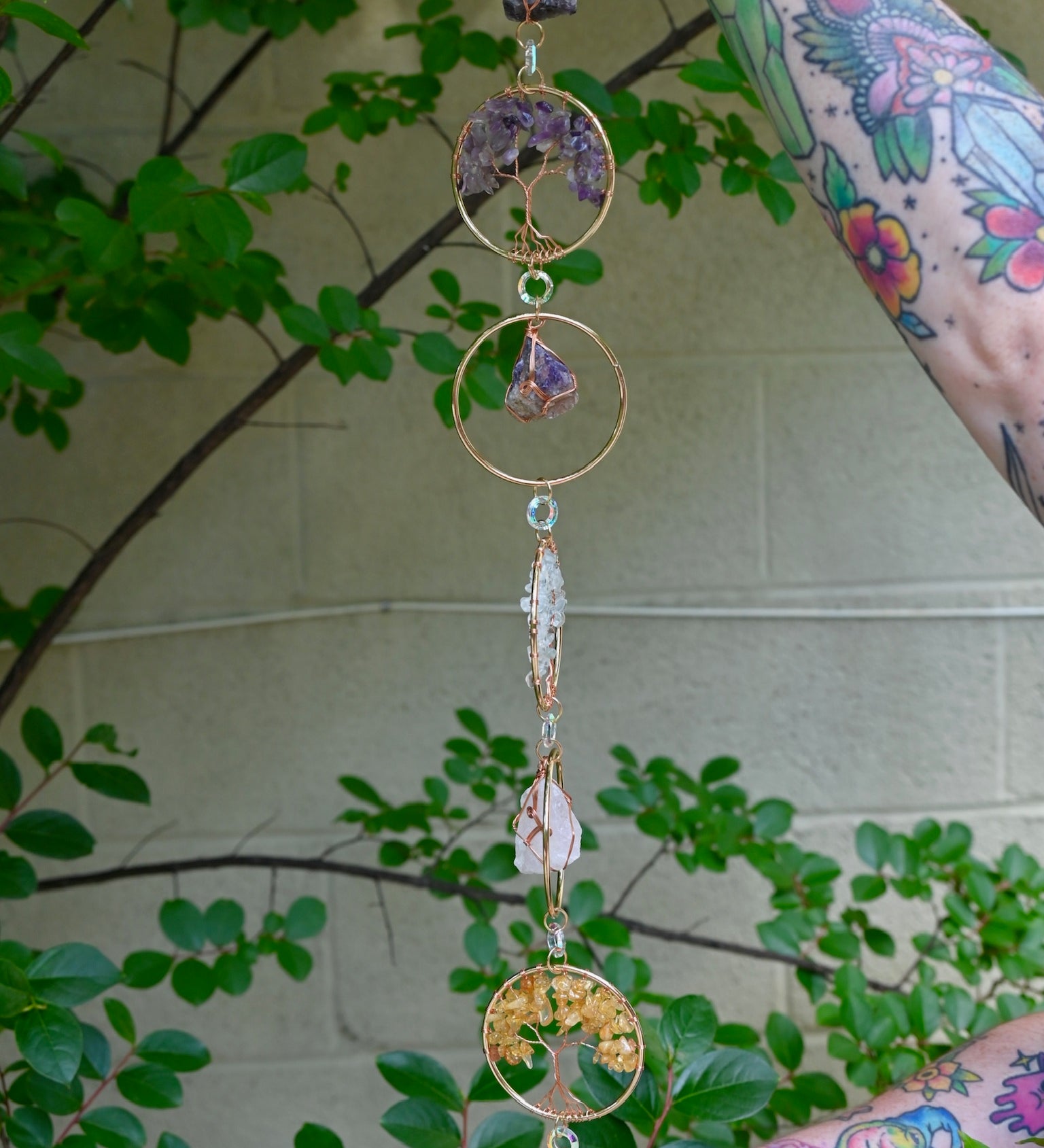 Someone holding the Chakra Gemstone Suncatcher with Amethyst, Citrine, and Quartz in front of a cream-colored brick wall with greenery.