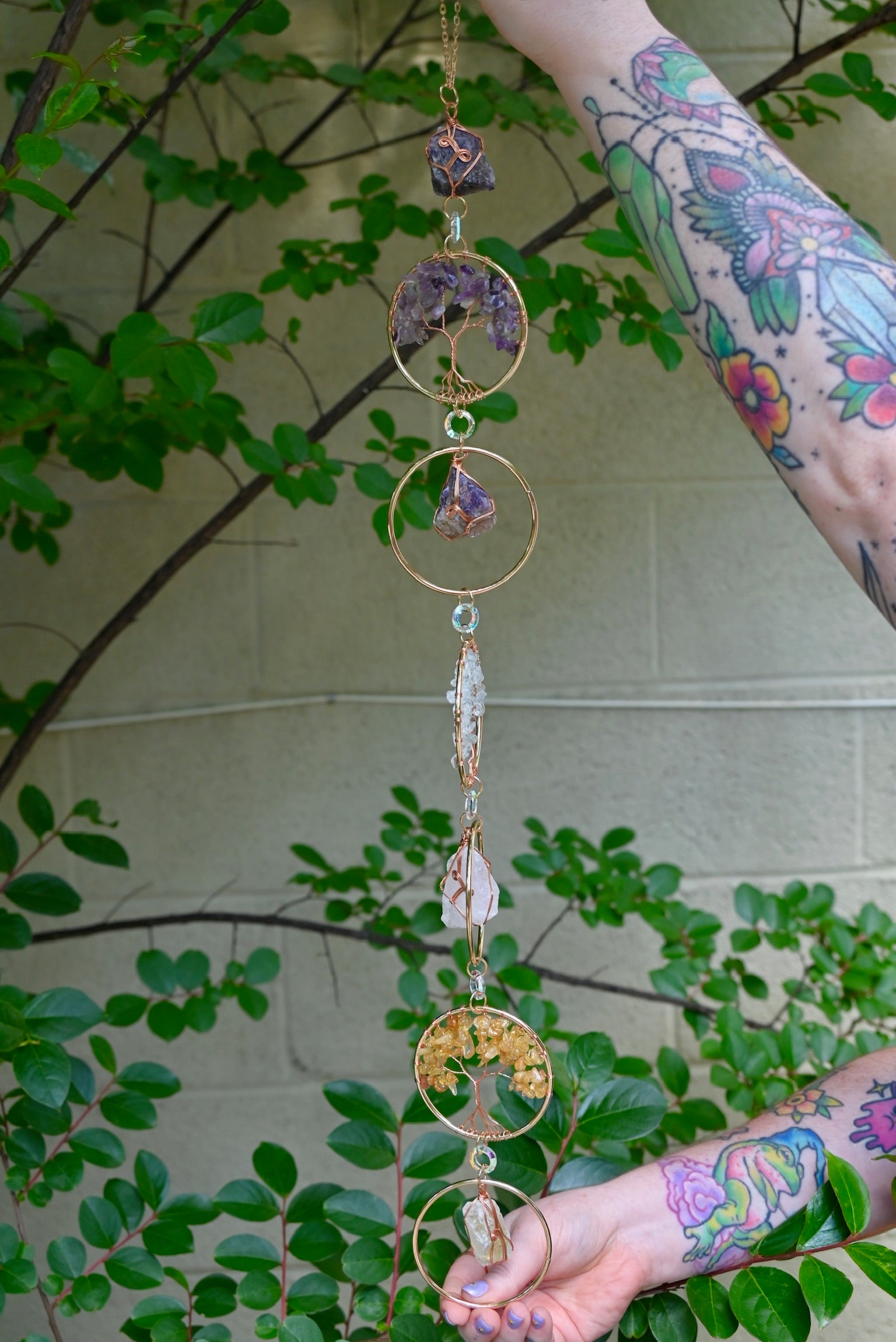 Someone holding the Chakra Gemstone Suncatcher with Amethyst, Citrine, and Quartz in front of a cream-colored brick wall with greenery.