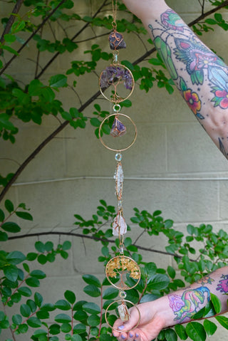 Someone holding the Chakra Gemstone Suncatcher with Amethyst, Citrine, and Quartz in front of a cream-colored brick wall with greenery.