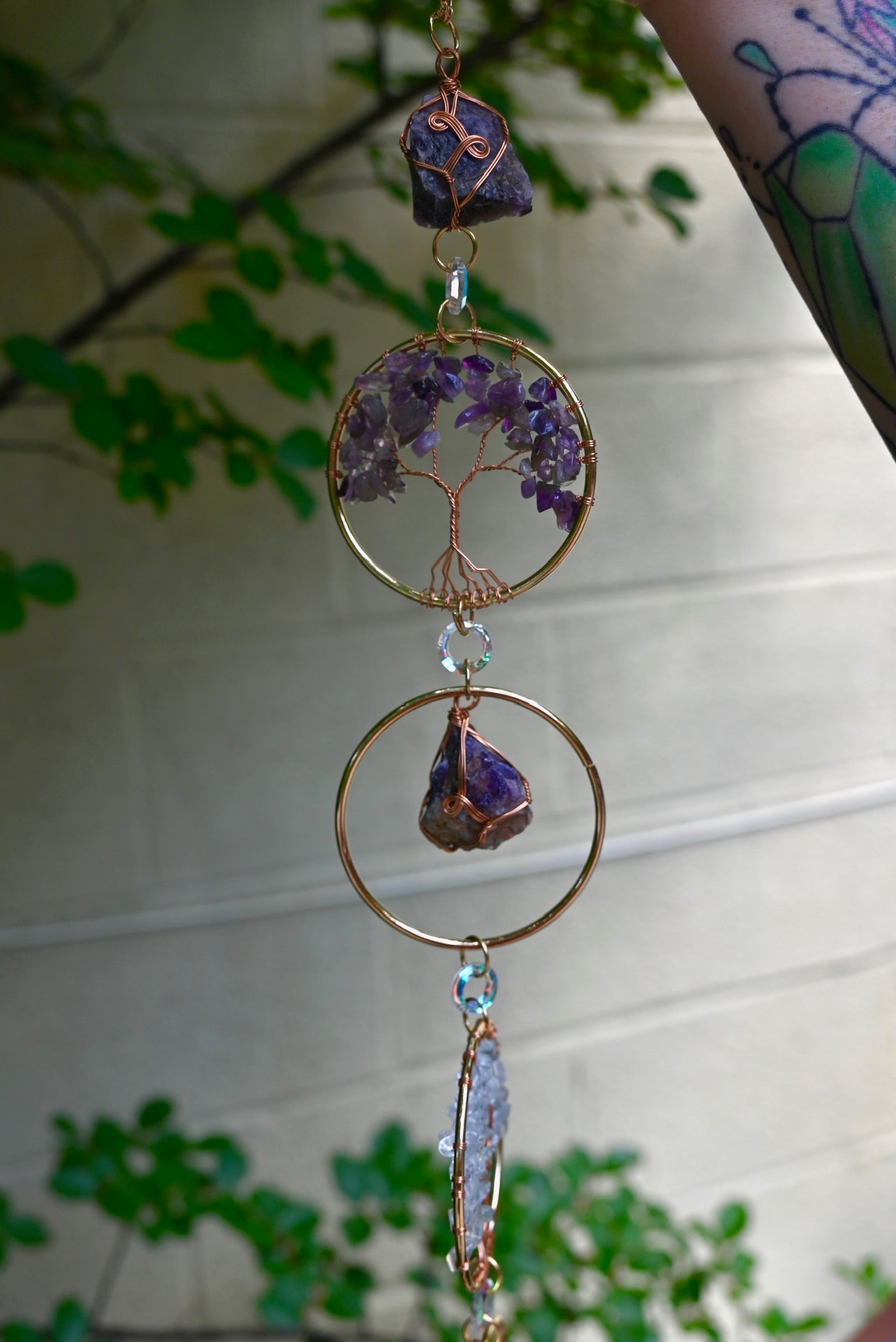Someone holding the Chakra Gemstone Suncatcher with Amethyst, Citrine, and Quartz in front of a cream-colored brick wall with greenery.