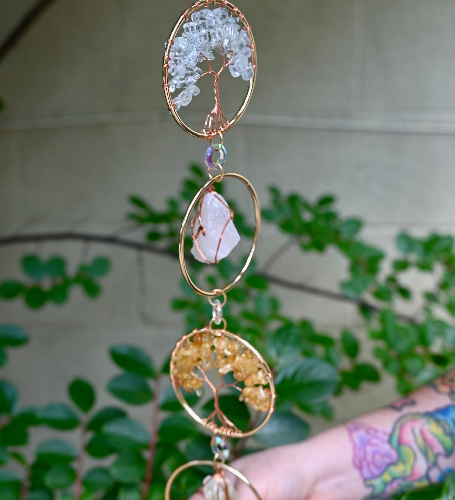 Someone holding the Chakra Gemstone Suncatcher with Amethyst, Citrine, and Quartz in front of a cream-colored brick wall with greenery.