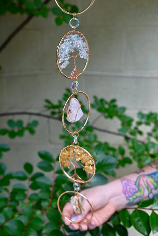 Someone holding the Chakra Gemstone Suncatcher with Amethyst, Citrine, and Quartz in front of a cream-colored brick wall with greenery.