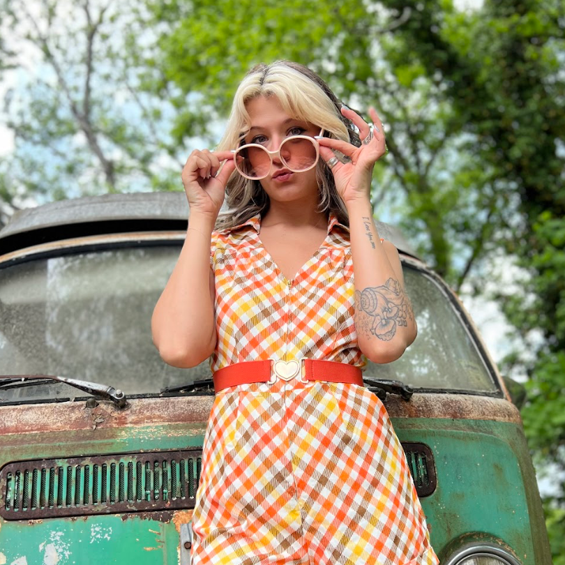 Woman in a retro disco jumpsuit standing in front of a vintage vehicle with trees in the background