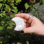 Hand holding a semi-transparent flower agate crystal moon with a natural background of greenery and flowers