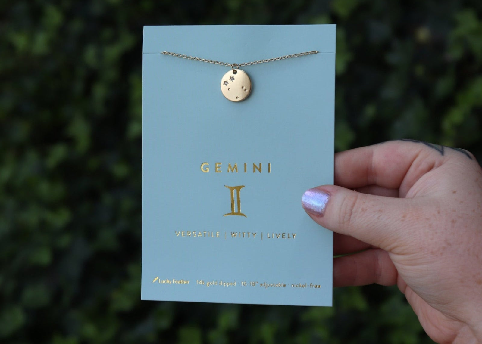 Someone holding the Gemini 14k Gold Dipped Nickel-Free Zodiac Necklace on its card in front of greenery.