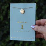 Someone holding the Gemini 14k Gold Dipped Nickel-Free Zodiac Necklace on its card in front of greenery.