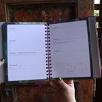 Someone holding the 2026 Lunar and Seasonal Northern Hemisphere Planner Calendar open in front of a wooden door, showing off the pages.
