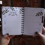 Someone holding the Woodland Wardens 2026 Weekly Planner in front of a door, showing off the pages.