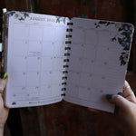 Someone holding the Woodland Wardens 2026 Weekly Planner in front of a door, showing off the pages.