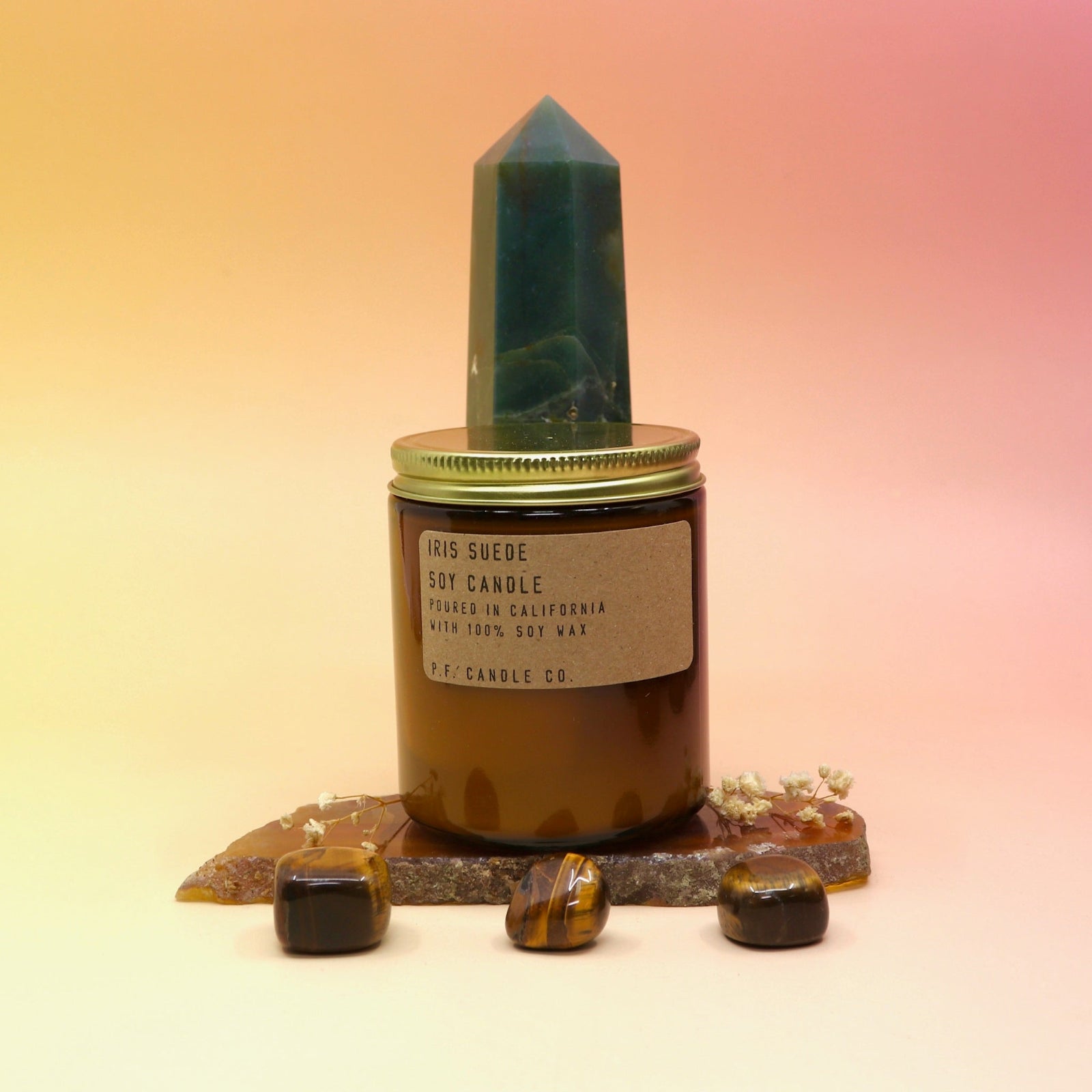 The Iris Suede Standard Soy-Wax Candle on an agate slice and in front of a crystal tower all on a pink and yellow background.