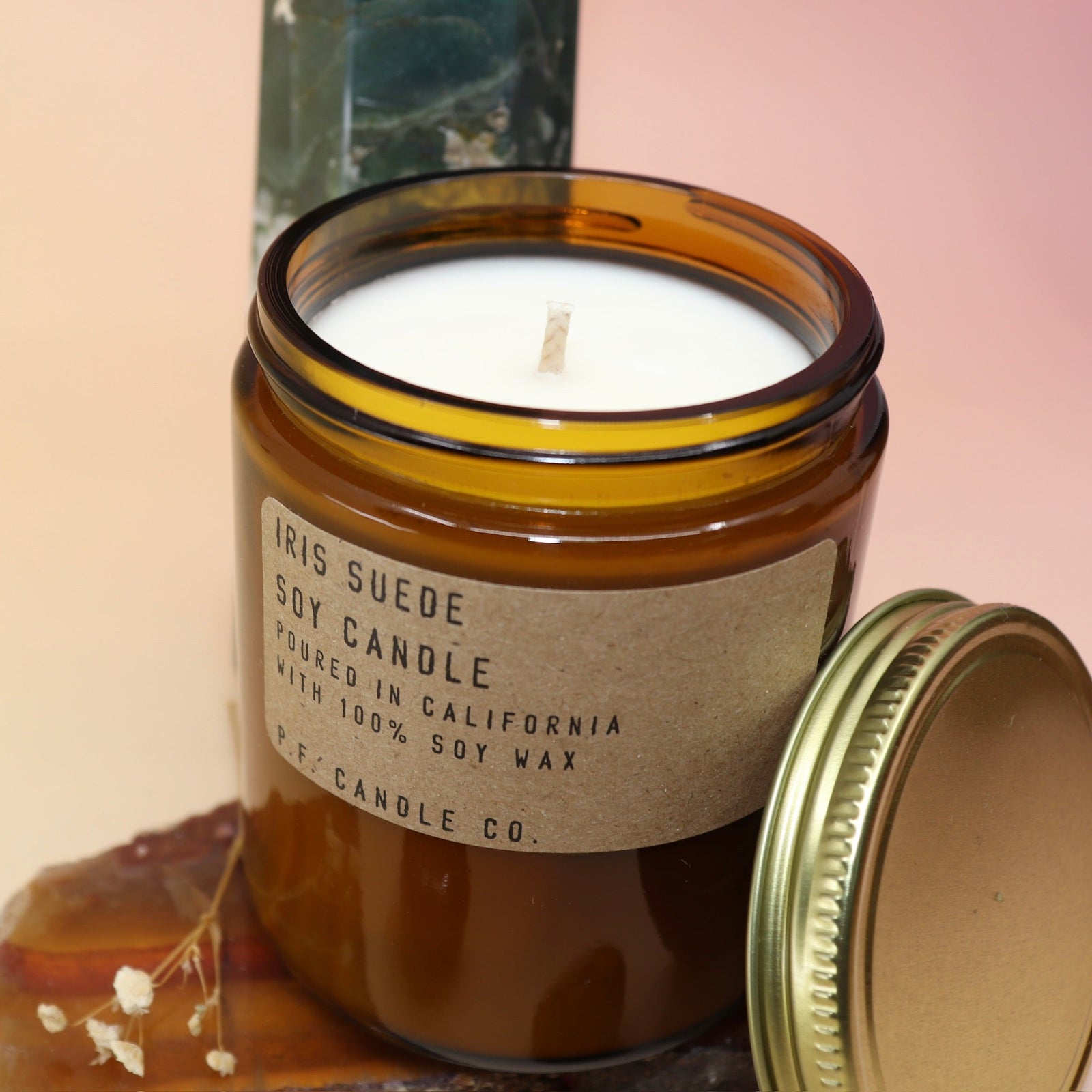 An up-close image of the Iris Suede Standard Soy-Wax Candle on an agate slice and in front of a crystal tower all on a pink and yellow background.
